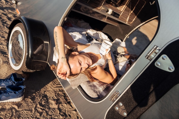 Cup of coffee. Blonde-haired handsome man lying in his compact trailer having some cup of coffee