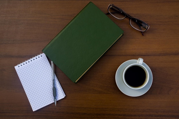 Cup of coffee, book, notepad, pen and eyeglasses on wooden desk