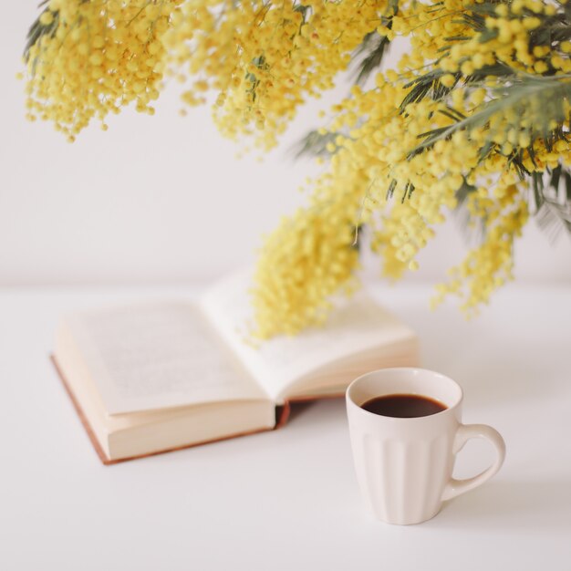 Cup of coffee and a book on white desk with yellow flowers bouquet mimosa.