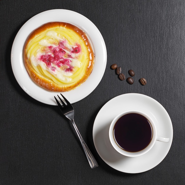 Cup of coffee and bun with custard and berries on black stone background