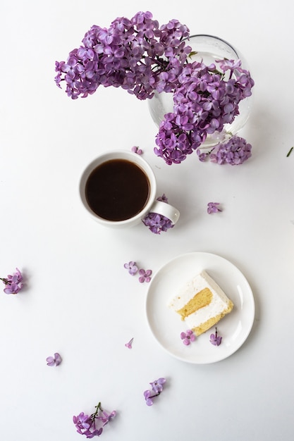Чашка кофе и рожки для торта из натюрморта с букетом сирени на белом столе, чашка кофе, тарелка с куском торта. Международный женский день, 8 марта