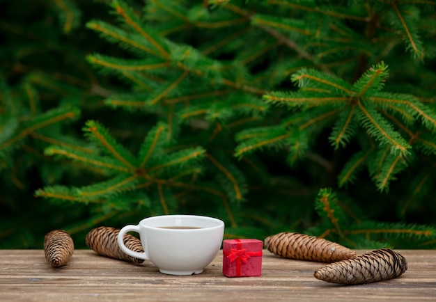 Cup of coffee and Christmas gift on wooden table with spruce branches on background