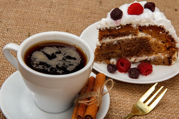 Cup of coffee, cinnamon, fork and slice of biscuit cake decorated with whipped cream and raspberries on table with sackcloth