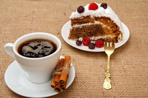 Cup of coffee, cinnamon, fork and slice of biscuit cake decorated with whipped cream and raspberries on table with sackcloth