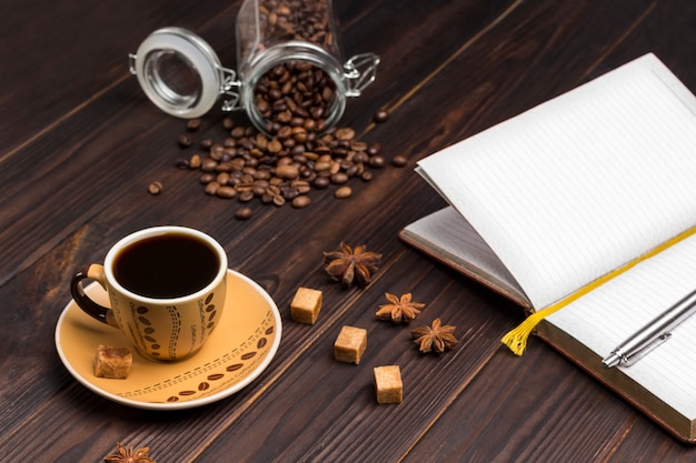 Cup of coffee, coffee beans in glass jar. Open notebook with pen. On table spices star anise and pieces of brown sugar.