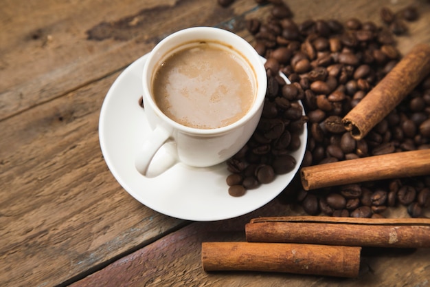 A cup of coffee, coffee seeds, cinnamon and a book on a wooden table