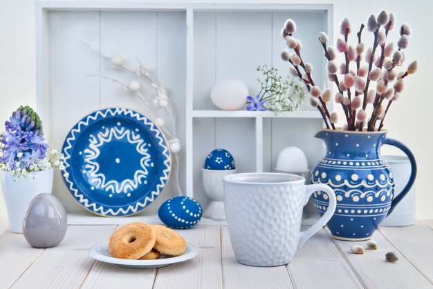 Cup of coffee and cookies on white table with bunch of pussy-willow in blue ceramic jar