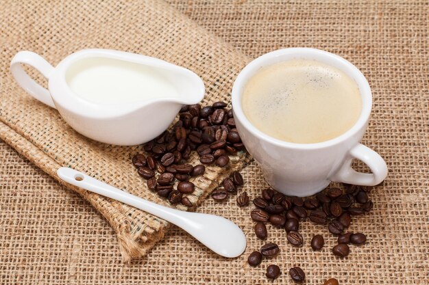 Cup of coffee, cream sauceboat, spoon, coffee beans and canvas sack on sackcloth. Top view.