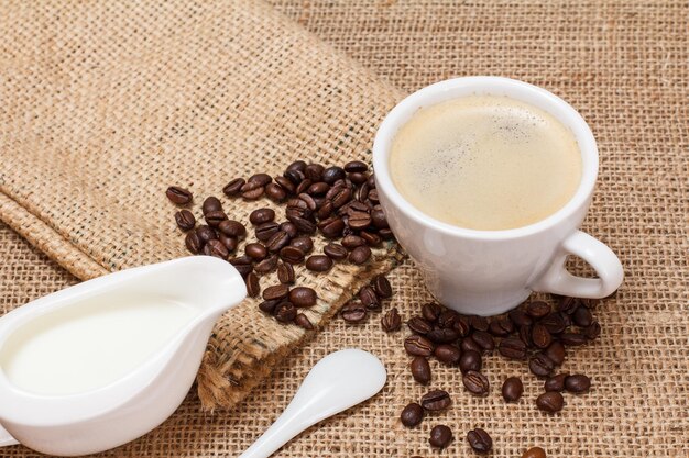 Чашка кофе со сливками, соусником, ложкой, кофейными зернами и холщовым мешком на мешковине, вид сверху