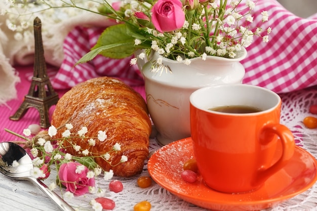 Cup of coffee and croissant are decorated by the small Eiffel Tower, napkins, roses and candies on a white wooden table