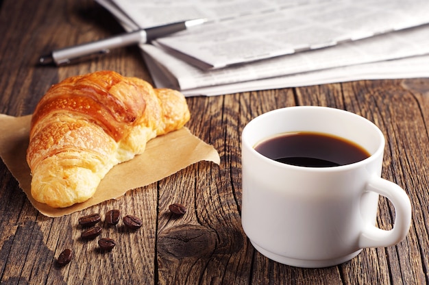 Cup of coffee, croissant and newspaper on wooden table