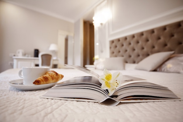 커피 한 잔, 크루아상 및 펼친 책은 밝고 아늑한 방에 침대에 놓여 있습니다.