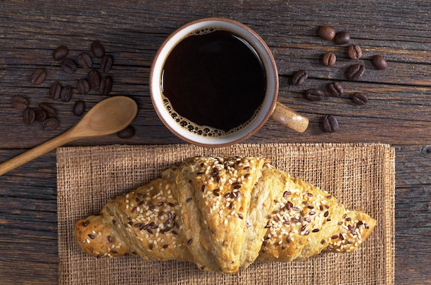 Cup of coffee and croissant with sesame seeds for breakfast on old wooden table, top view