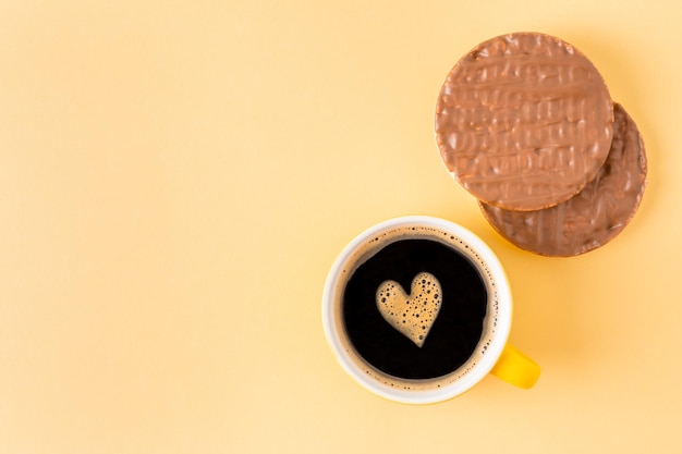 Foto tazza di caffè decorata con cuore di schiuma vicino a torte di riso ricoperte di cioccolato su sfondo giallo, vista dall'alto, spazio vuoto