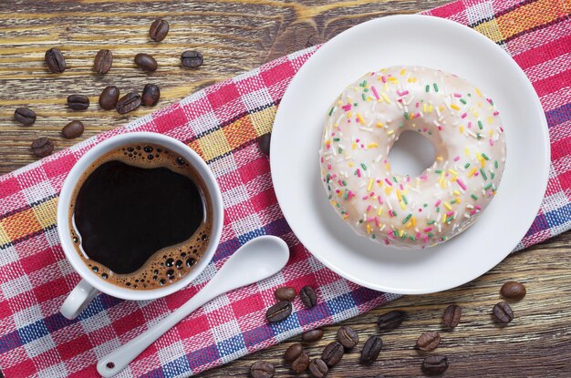 Cup of coffee and delicious donut with frosting and decorated colorful sprinkles on red napkin is located on an old wooden table, top view