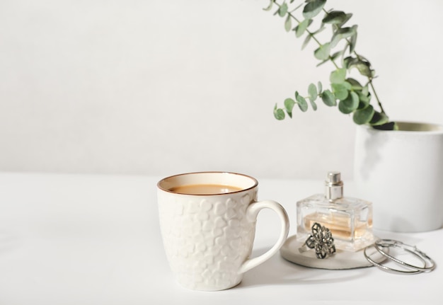 A cup of coffee and female accessories on a table a bottle of\
perfume and jewellery and green leaves on white table good morning\
concept