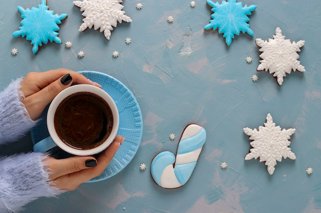 Tazza di caffè in mani femminili e biscotti di panpepato a forma di fiocchi di neve su azzurro