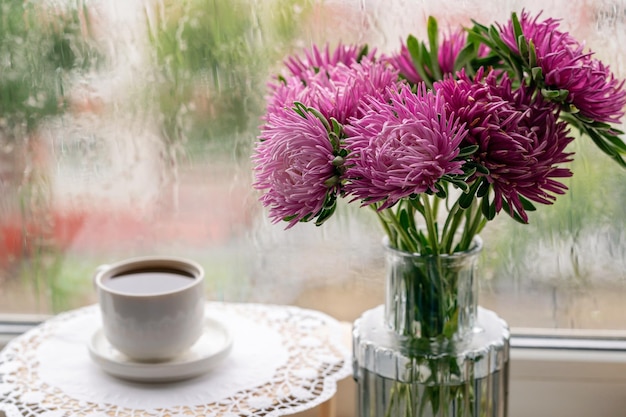 A cup of coffee and flowers in the vase standing next to a\
rainy window.