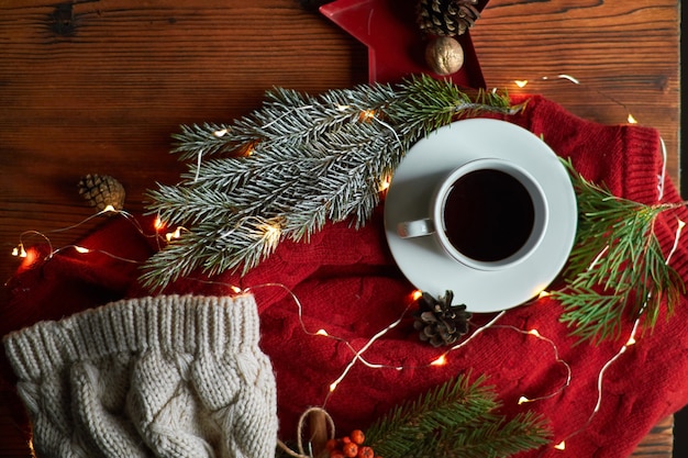 A cup of coffee and a garland on a wooden tray with a cozy red sweater and a warm hat. New Year's still life with a fir branch and mountain ash