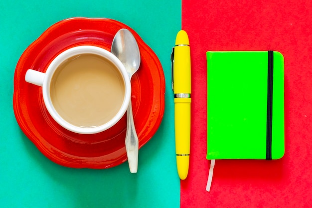 Чашка кофе рядом с зеленой записной книжкой и желтой авторучкой, все готово, чтобы делать заметки или организовывать день после завтрака.