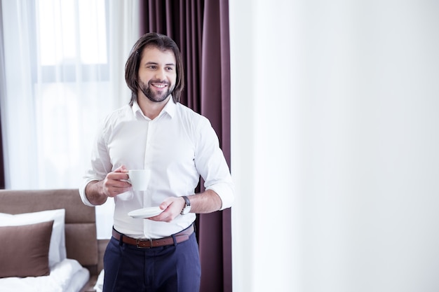 A cup of coffee. Joyful positive businessman smiling while enjoying his morning drink