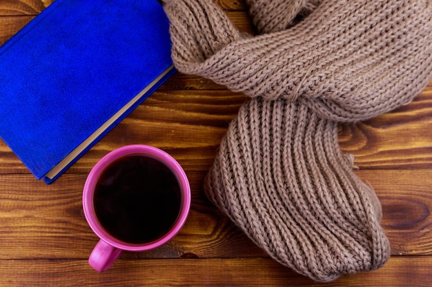 Cup of coffee, knitted scarf and book on wooden background