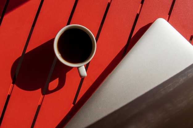 A cup of coffee and a laptop on a red table kind of light hard
light shadows top view