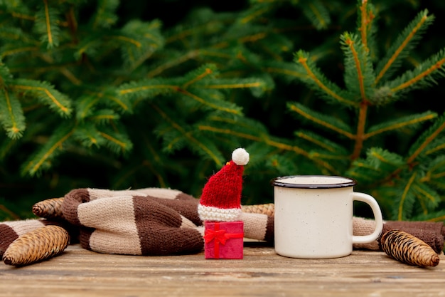 Cup of coffee and little Christmas gift in hat on wooden table with spruce branches on background