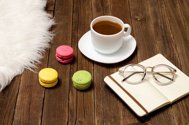 Cup of coffee, macarons, a notebook and glasses on a wooden table