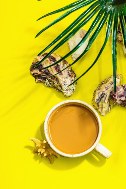 A cup of coffee in a marine style starfish oysters palm leaves hard light dark shadow bright yellow background flat lay top view