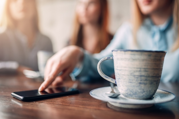 一杯のコーヒーと木製のテーブル、クローズアップの携帯電話。カフェで女の子のレジャー。ソファの上のレストランに座ってコーヒーを飲み、おしゃべり会議の女友達