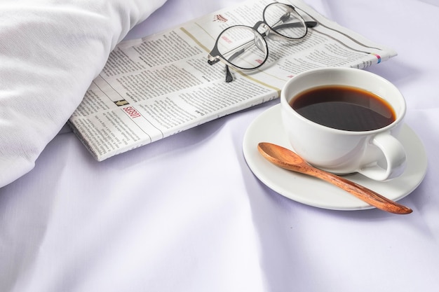 A cup of coffee and a newspaper on a white bed in the morning.