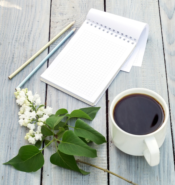 Cup of coffee, notepad and a lilac branch on the white wooden table