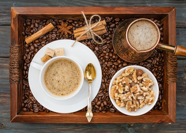 Cup of coffee, nuts in the bowl, coffeepot, cinnamon, anise, sugar, coffee beens on a wooden tray