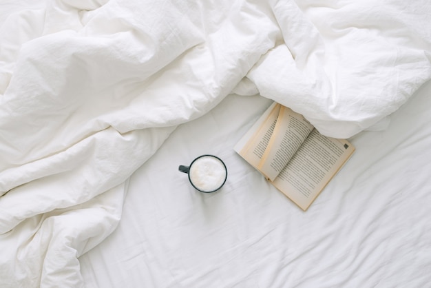 A cup of coffee and an open book lie on a white open bed. Top view