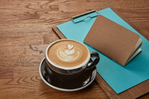 Cup of coffee represented on wooden table with notebook and clipboard near. Delicious cappuccino, coffee with milk.