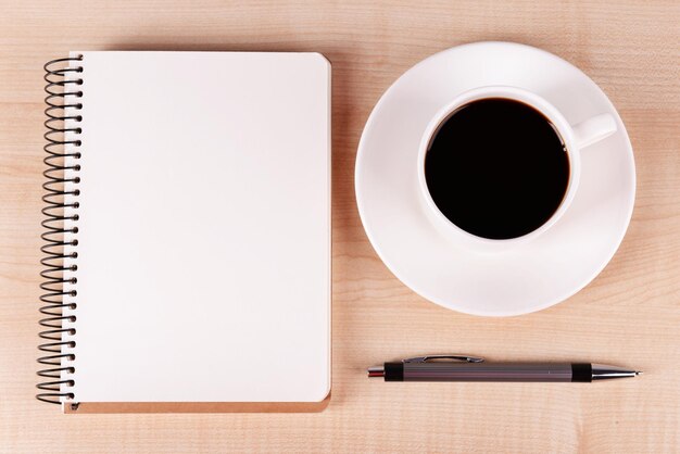 Cup of coffee on saucer with notebook and pen on wooden table background