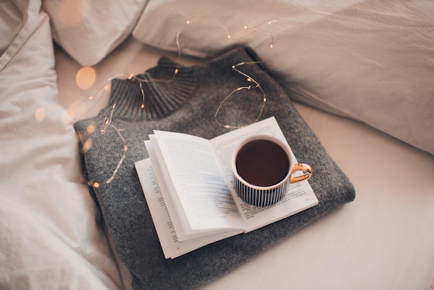 Cup of coffee stay on knitted textile sweater in bed with paper open book with christmas glowing lights close up