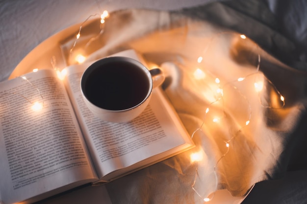 Cup of coffee stay on open paper book on tray with glowing lights close up. Top view. Reading and working at night.