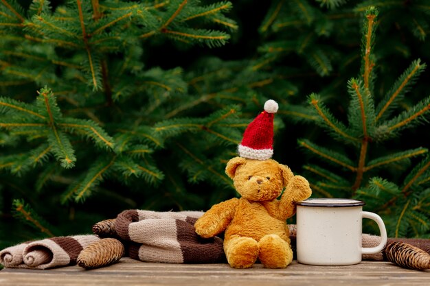 Cup of coffee and teddy bear in hat on table with spruce branches on background