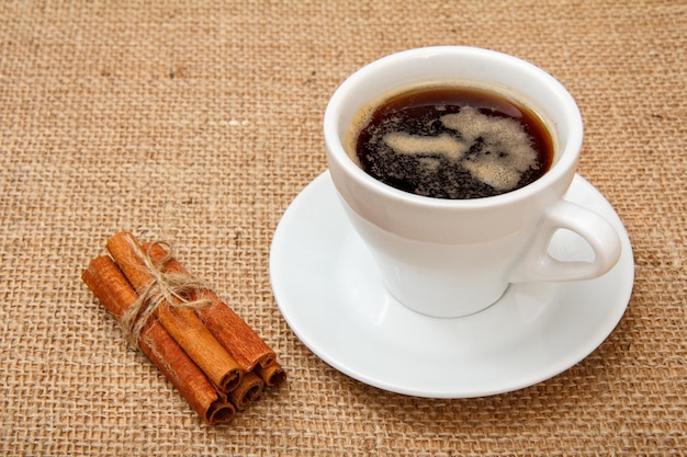 Cup of coffee on white saucer and cinnamon on table with sackcloth.