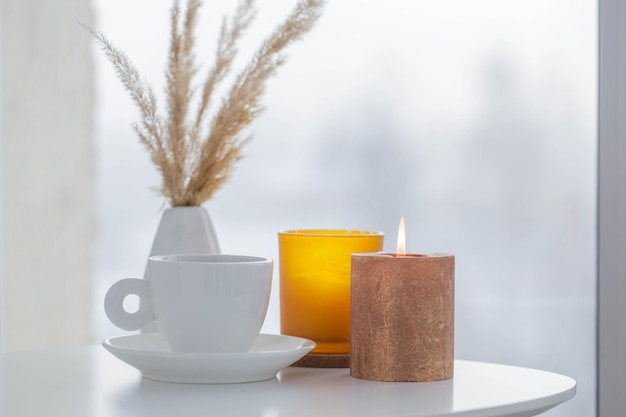 cup of coffee with burning candles  on white table by window on winter