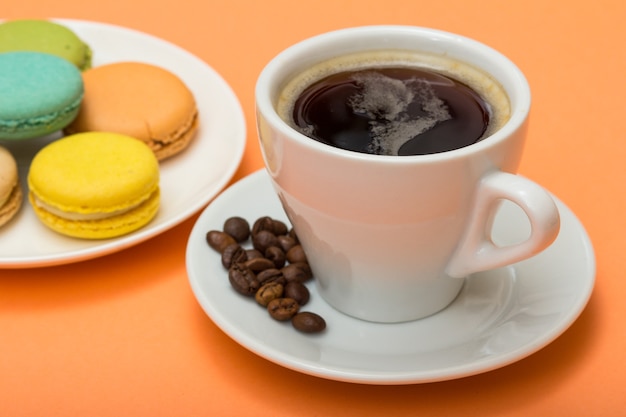 Cup of coffee with coffee beans and delicious macarons cakes of different color on white porcelain plate with peach colored background. Selective focus on cup