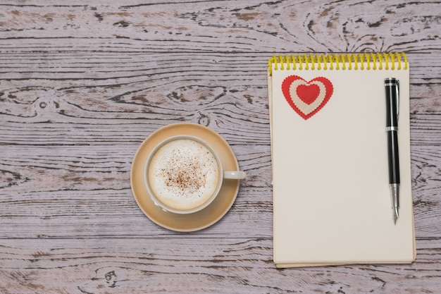 A Cup of coffee with cream and cinnamon, a notebook and a fountain pen on a wooden table