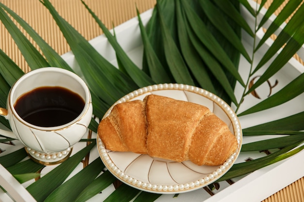 Cup of coffee with croissant on white tray with fern leaf in morning. Healthy food