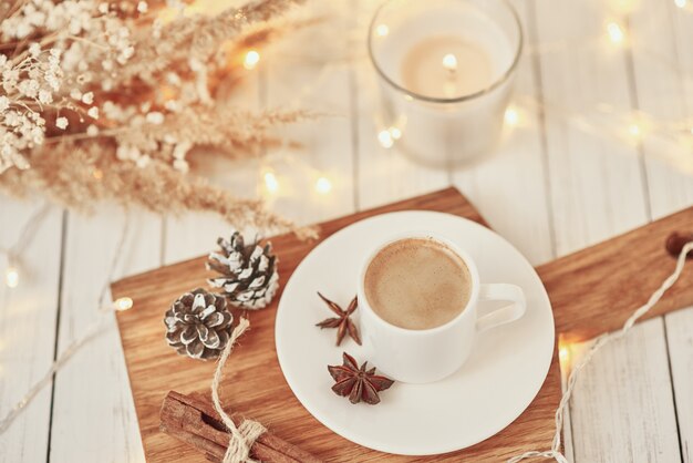 Cup of coffee with garland lights, burning candle and decoration on table. Cozy home concept