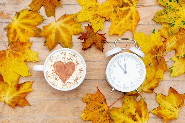 Cup of coffee with a heart of cinnamon and alarm clock on the table, maple leaves around