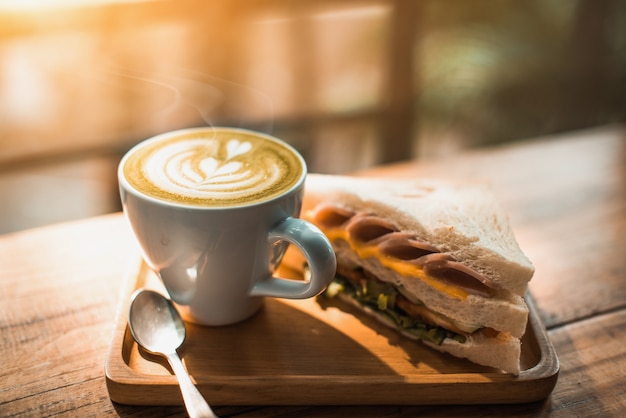 A cup of coffee with heart pattern in a white cup and Sandwich on wooden table background - Image