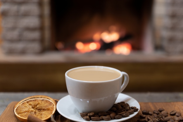 Cup of coffee with milk and coffee beans around, cinnamon sticks before cozy fireplace.