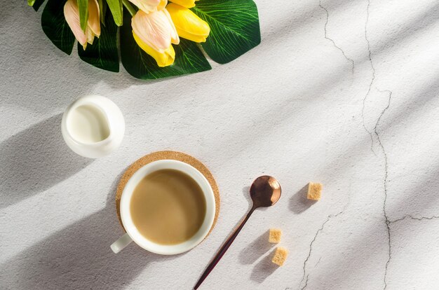 Cup of coffee with milk, a milk jug, brown sugar cubes and a bouquet of flowers on white table. Breakfast on a sunny day. Copy space.